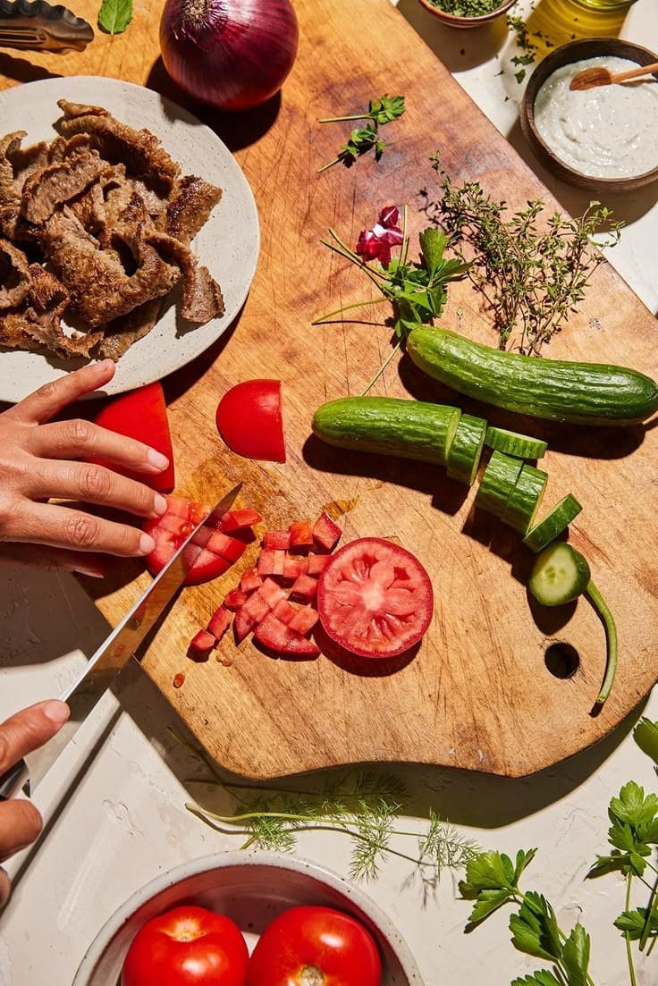 Chopping tomatoes and cucumber on a board beside grilled meat and fresh herbs